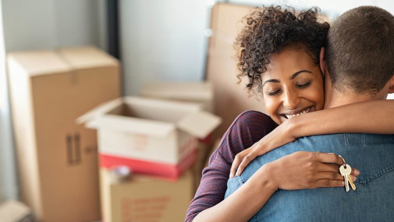 Young african woman holding home keys while hugging boyfriend in their new apartment after buying real estate. Lovely girl holding keys from new home and embracing man. Happy couple in their apartment around cardboard boxes.